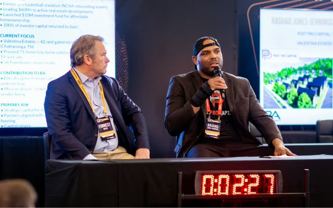 Rashad on stage with a microphone at a real estate panel, Valentina Estates rendering on the screen behind him