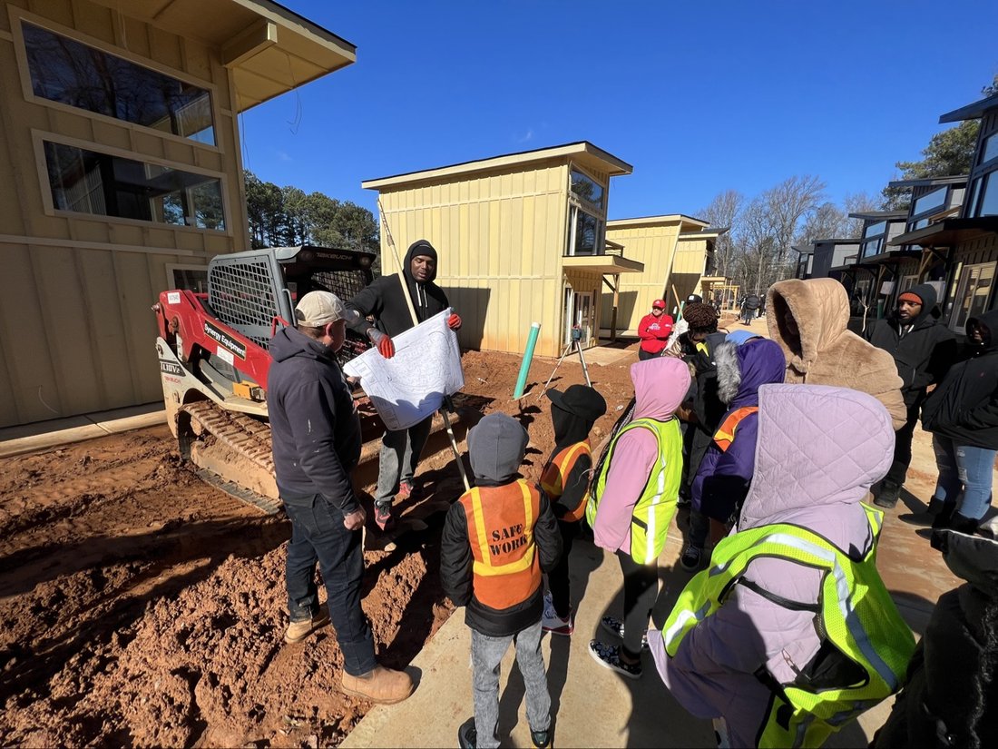 Kids in safety vests touring the tiny home site while a crew member holds plans by the equipment